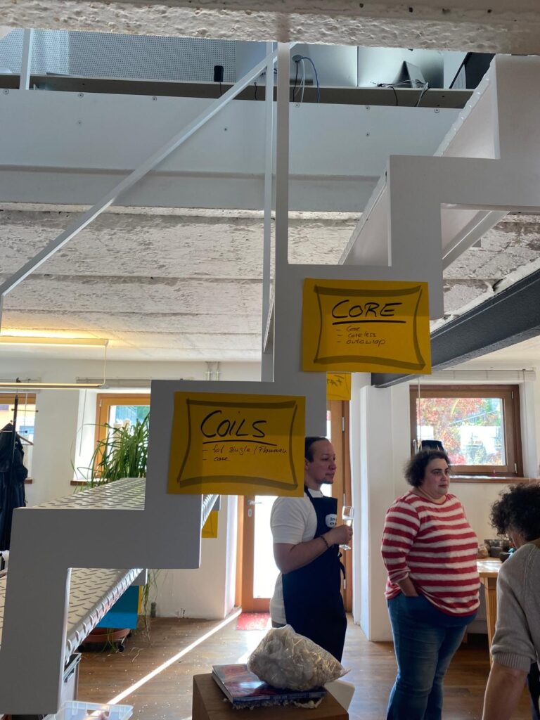 Industrial interior with white structures and yellow sign boards labeled CORE and COILS, people standing and talking in the background.