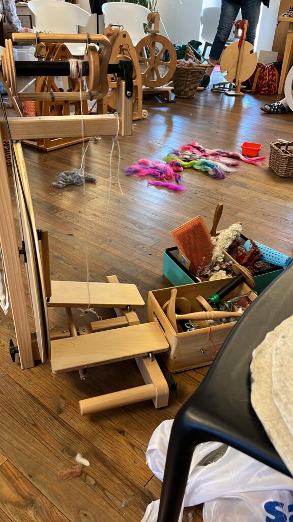 Weaving workshop scene with a large wooden loom in the foreground, colorful fiber rovings on the floor, and baskets of tools around a sunny studio space.