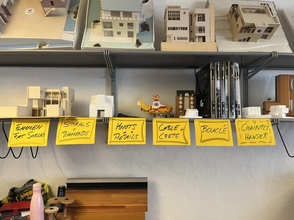 Row of architectural miniatures on a shelf above a desk, with yellow handwritten labels listing craft topics below.