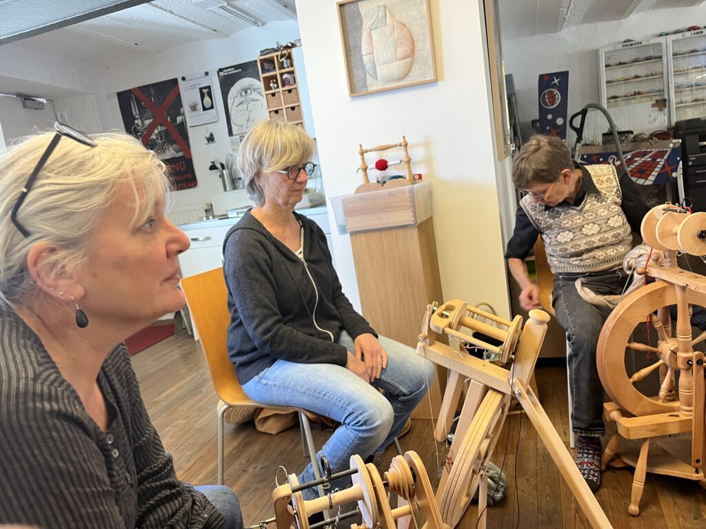 Three people sit in a craft room watching a man demonstrate weaving on a large wooden loom, yarn and supplies nearby.