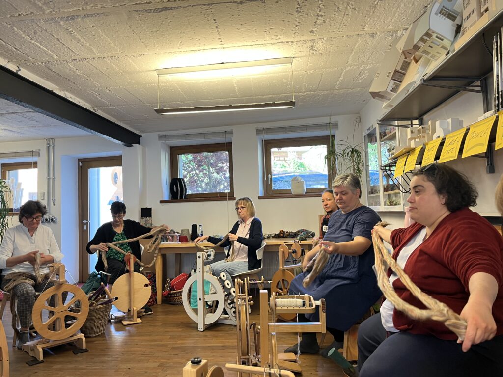 Adults seated around tables knitting with yarn and spinning wheels in a bright, cozy workshop with large windows and shelves.