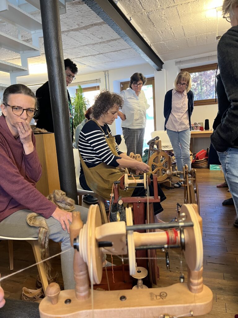 Group of adults in a craft workshop gathered around traditional spinning wheels; a woman in a striped shirt is actively spinning while others watch.
