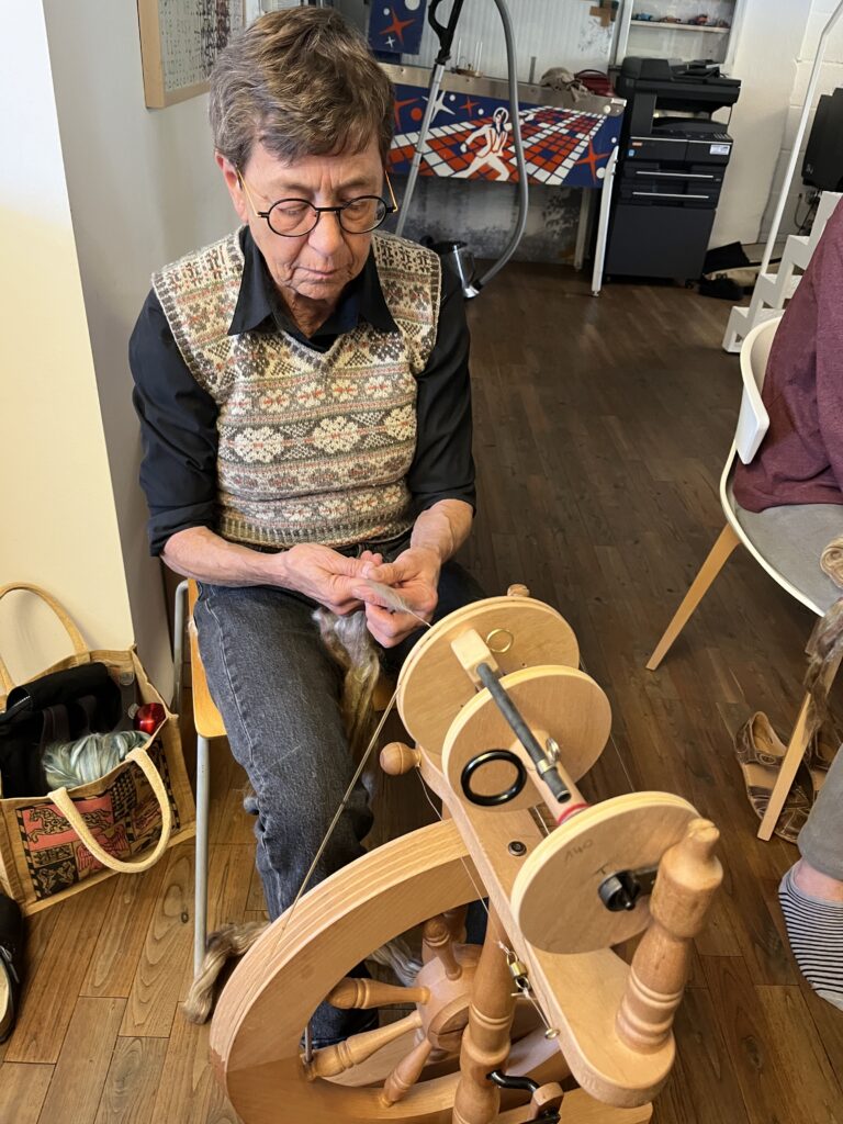 Older man with glasses sits at a spinning wheel, guiding wool yarn with his hands in a cozy workshop setting.