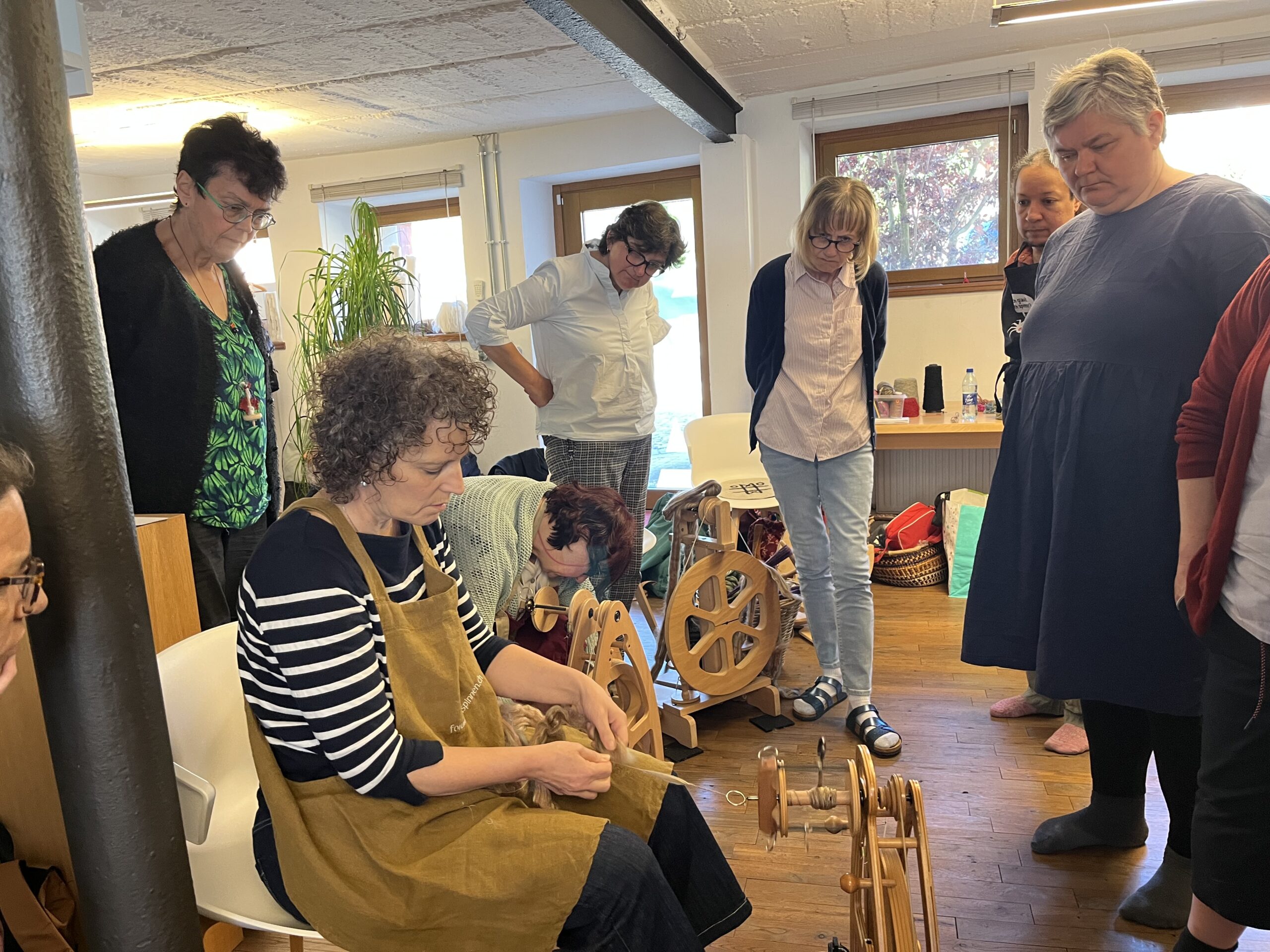 A woman sits at a loom weaving while others stand around watching in a bright workshop.