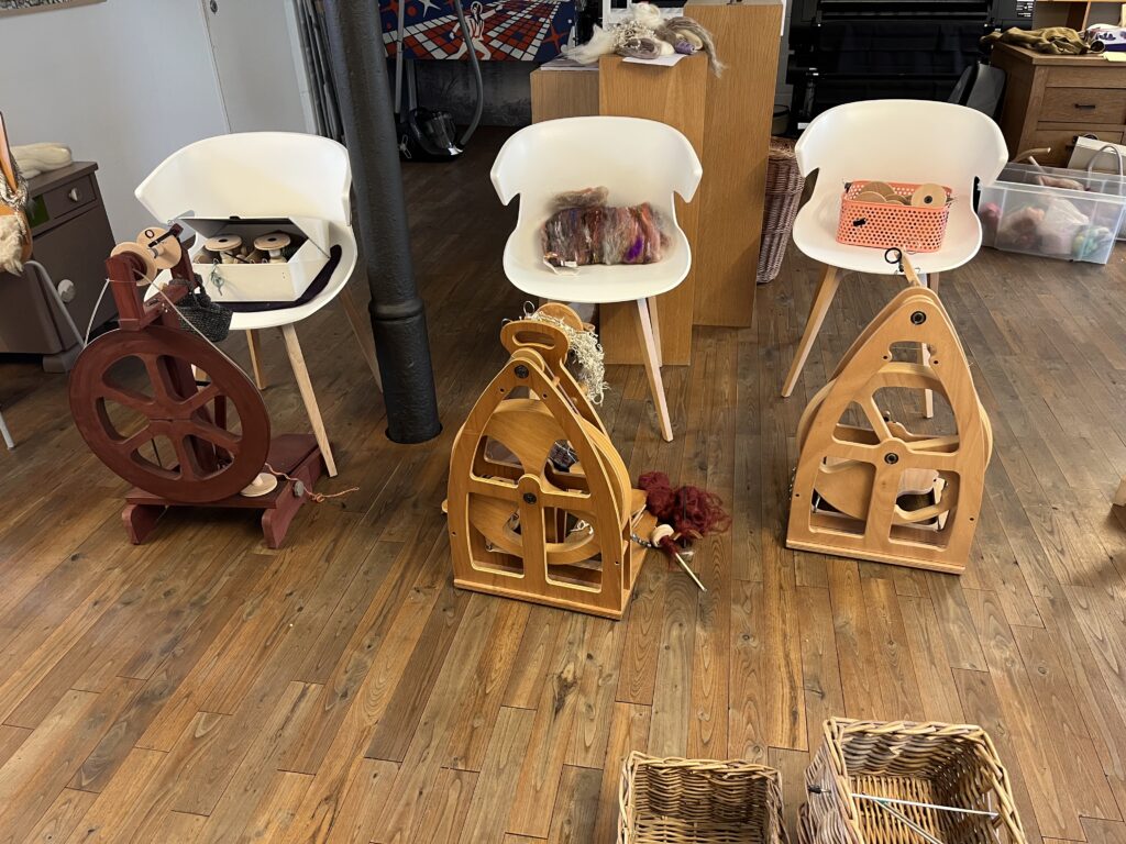 Fiber arts workspace with white chairs, spinning wheels, and baskets of yarn on a wooden floor.