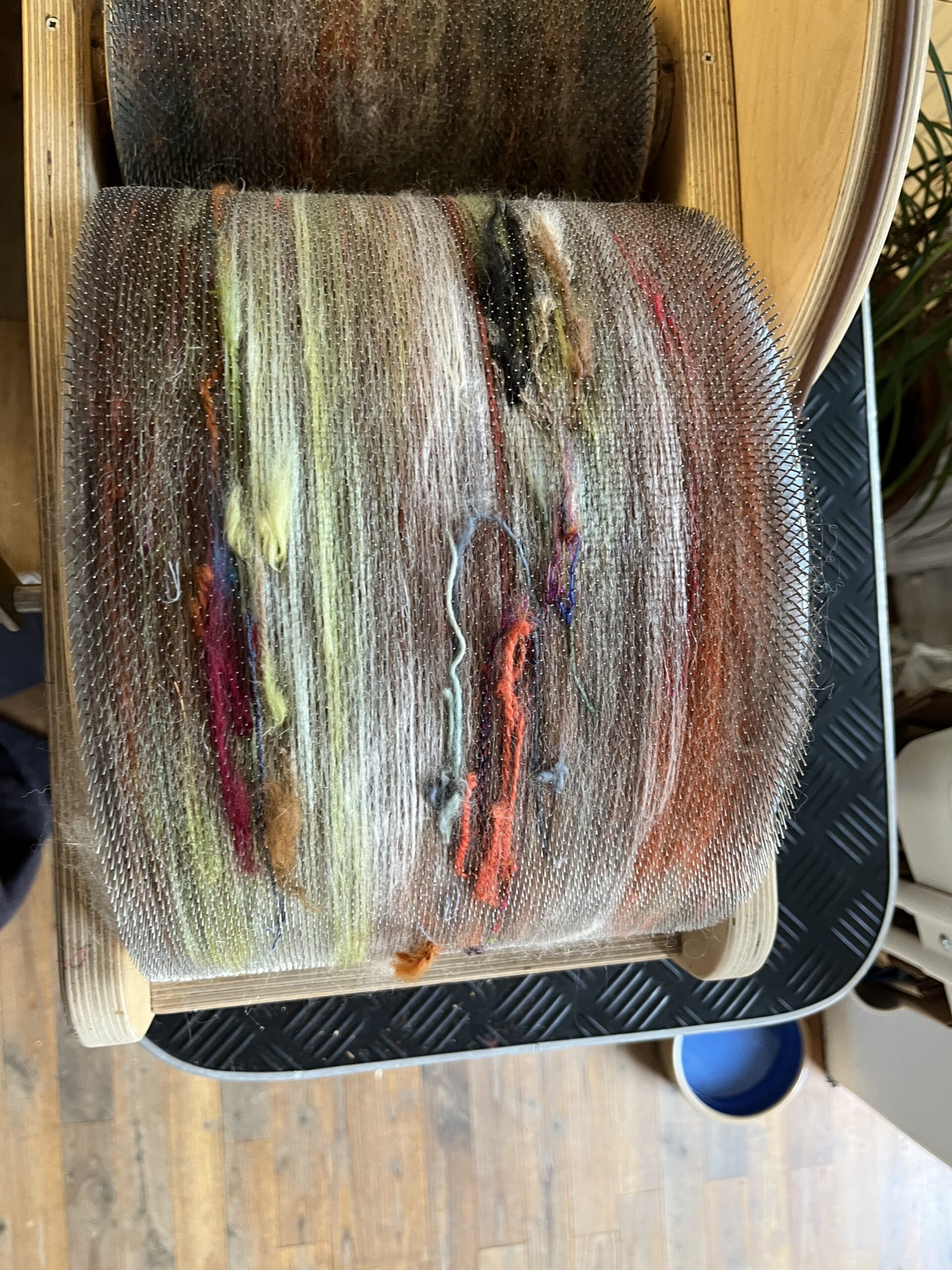 Close-up of a small weaving loom with multicolored yarns wound on the warp threads, showing rusty reds, greens, yellows and browns.