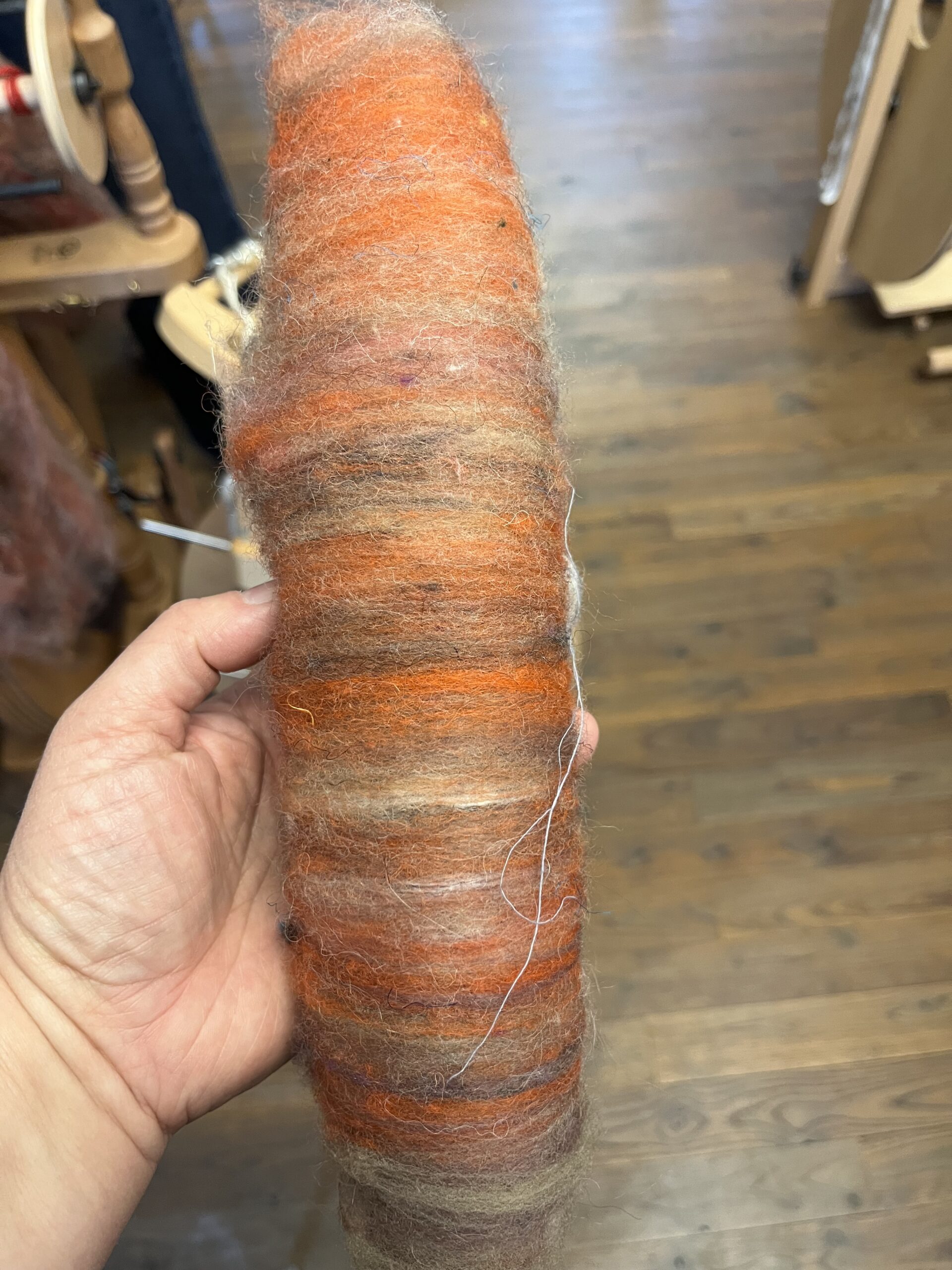 Close-up of a hand holding a thick, fuzzy skein of orange-brown variegated wool with striped bands; a spinning wheel is out of focus in the background.