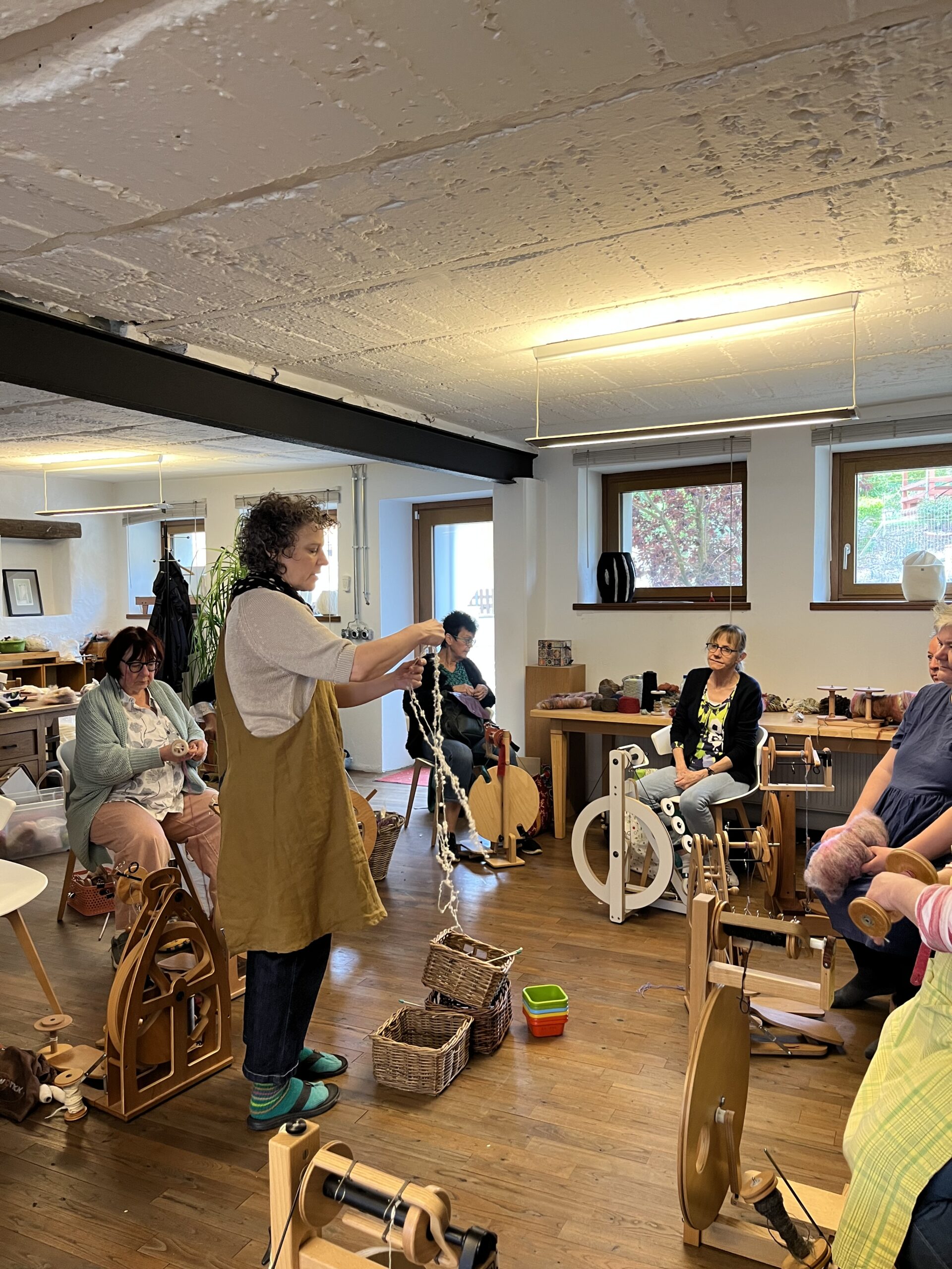 A group of people in a craft workshop using spinning wheels and weaving tools in a bright room with large windows.