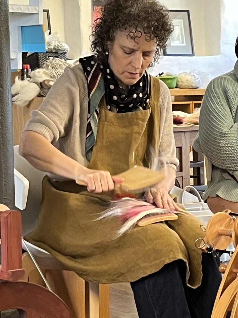Woman wearing a brown apron and polka-dot scarf cutting fabric at a craft workshop table.