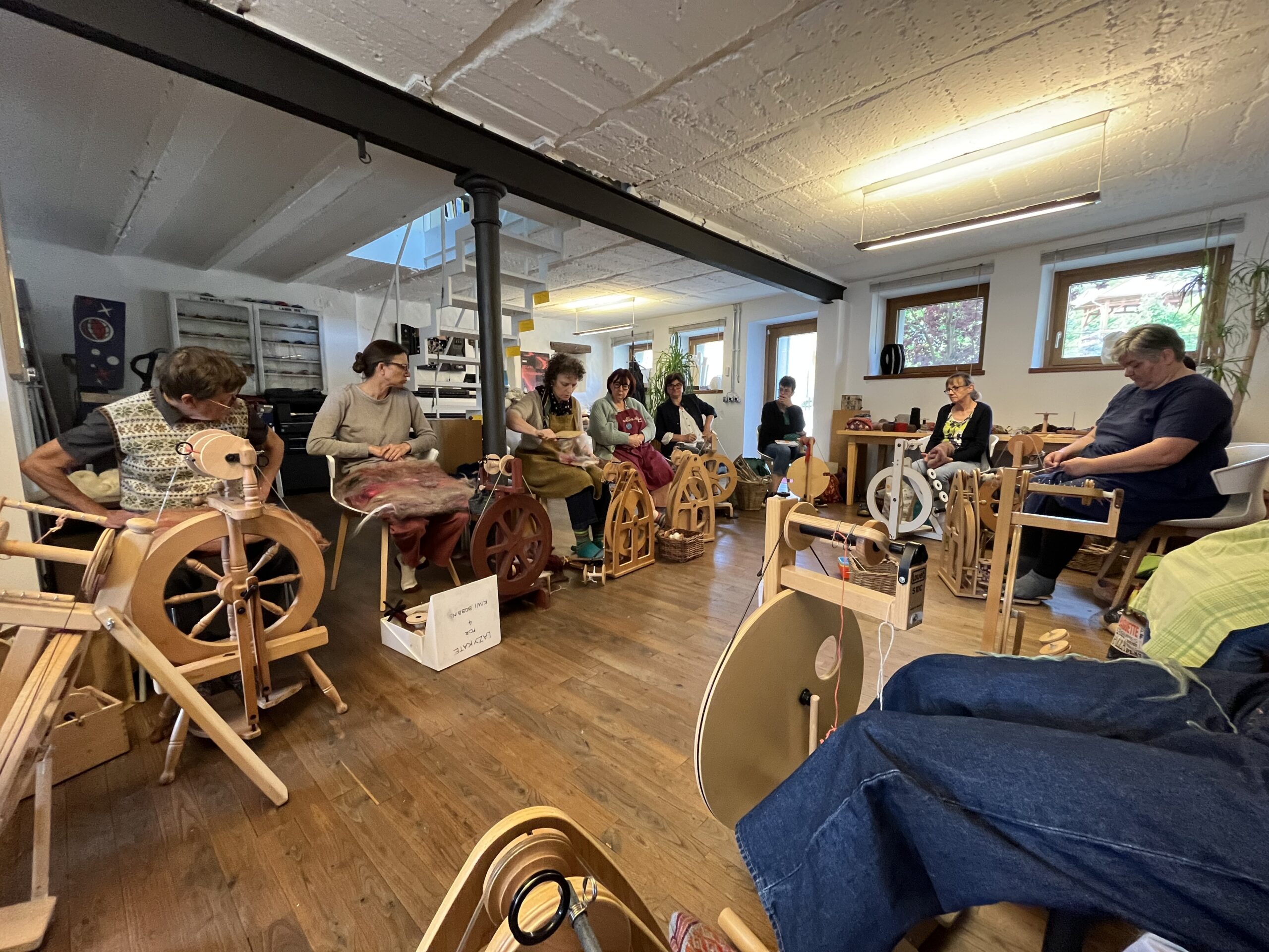 Group of adults in a bright studio, sitting in a row and weaving on spinning wheels during a workshop.