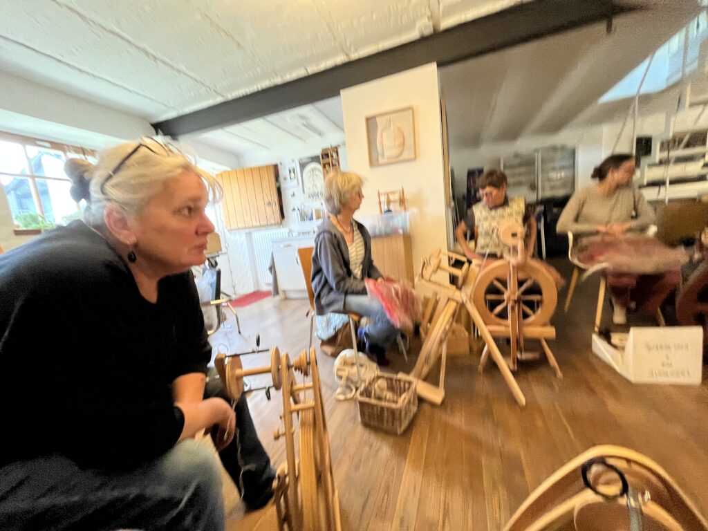 People gathered in a bright workshop with spinning wheels and craft tables visible in the foreground.