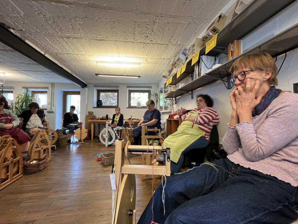 People seated in a craft workshop room with spinning looms and baskets, focused on projects under warm lighting.