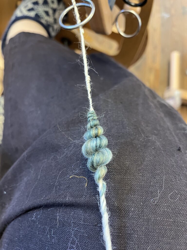 Close-up of a white cord being wrapped with teal and blue yarn into a spiral; crafting scissors and rings visible nearby.
