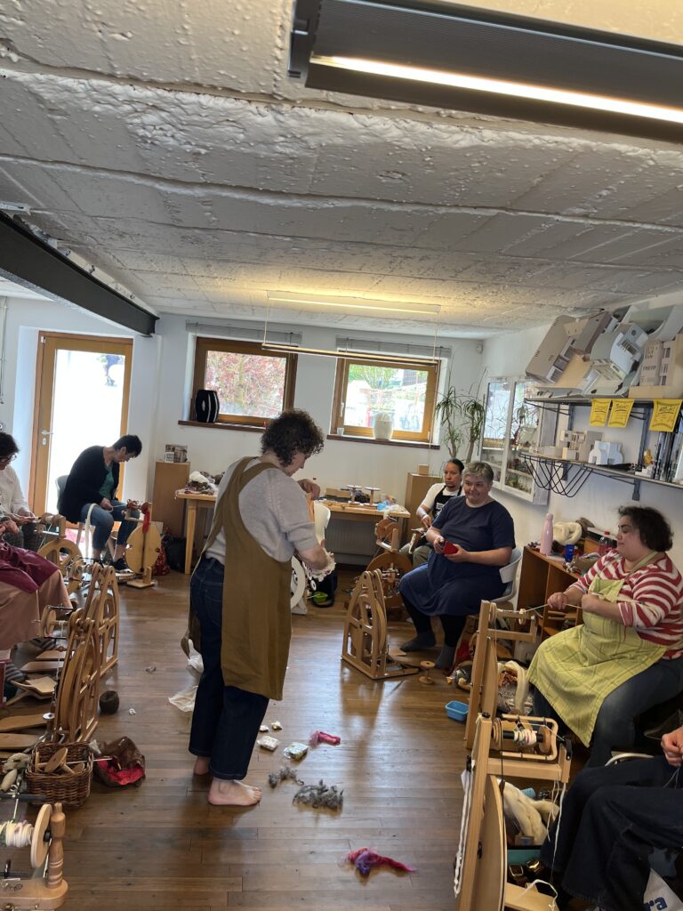 Group of people in a textile workshop weaving on wooden looms in a bright room with scattered yarns on the floor.