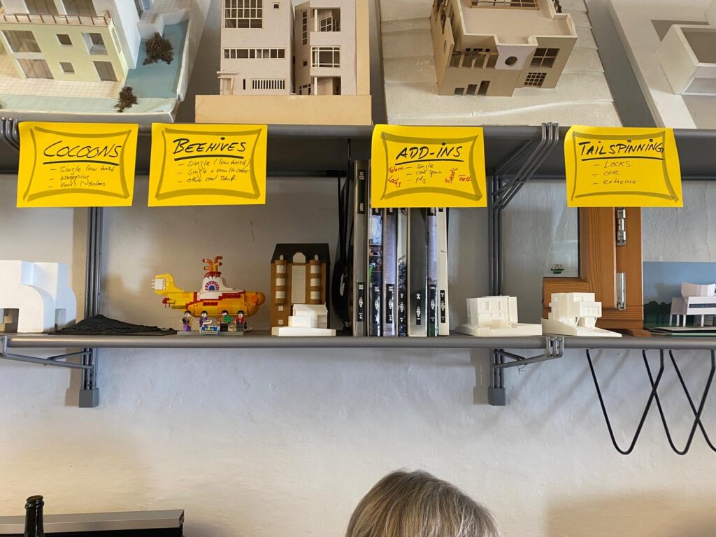 Row of bright yellow sticky notes with handwritten categories—Cocoons, Beehives, Add-Ins, Tailspinning—above a shelf with model buildings and books.