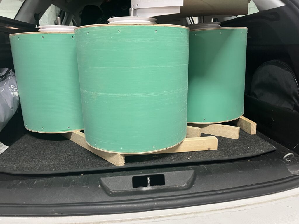 Green cylindrical drum containers stacked in a car trunk on wooden pallets, with white lids slightly open at the top.