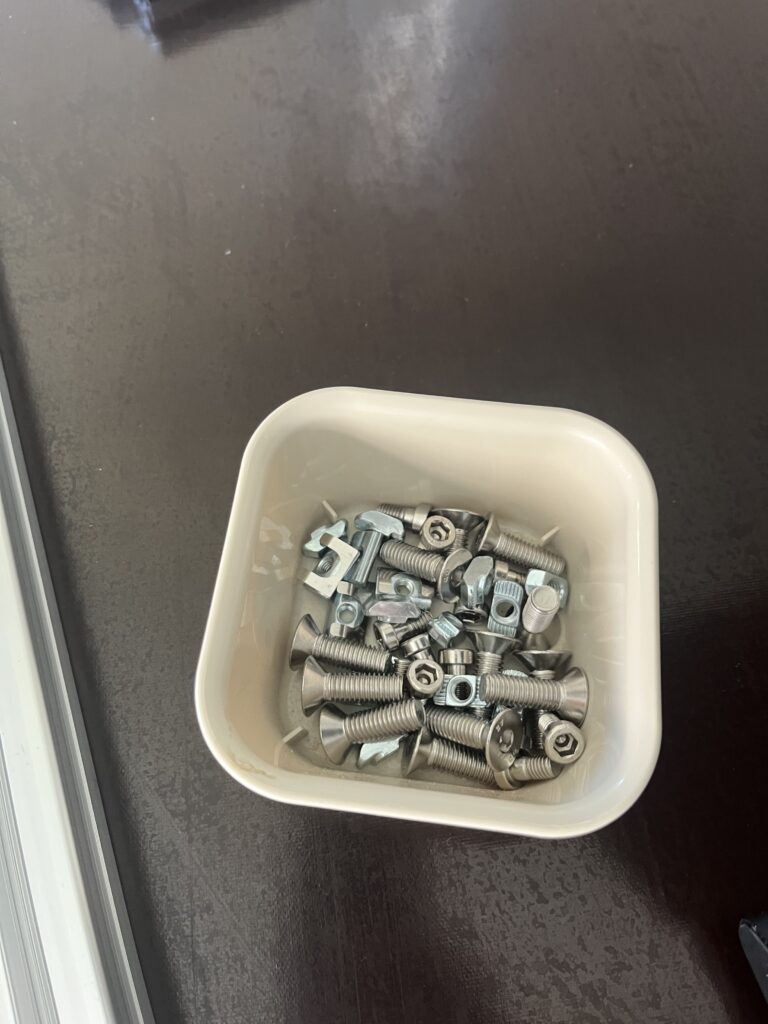 White plastic bin filled with assorted metal bolts, nuts, and washers on a dark floor