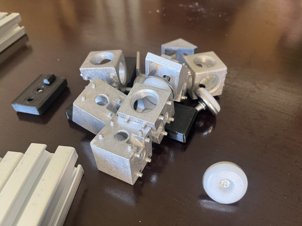 Pile of gray metal modular blocks with circular holes and a couple of small wheels on a dark table surface.