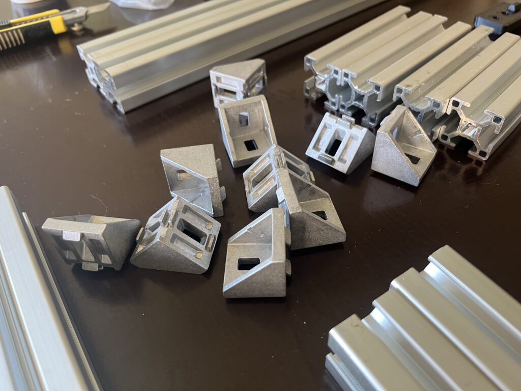 Assorted aluminum extrusion profiles and metal brackets laid out on a dark workshop table, with a utility knife in the background.