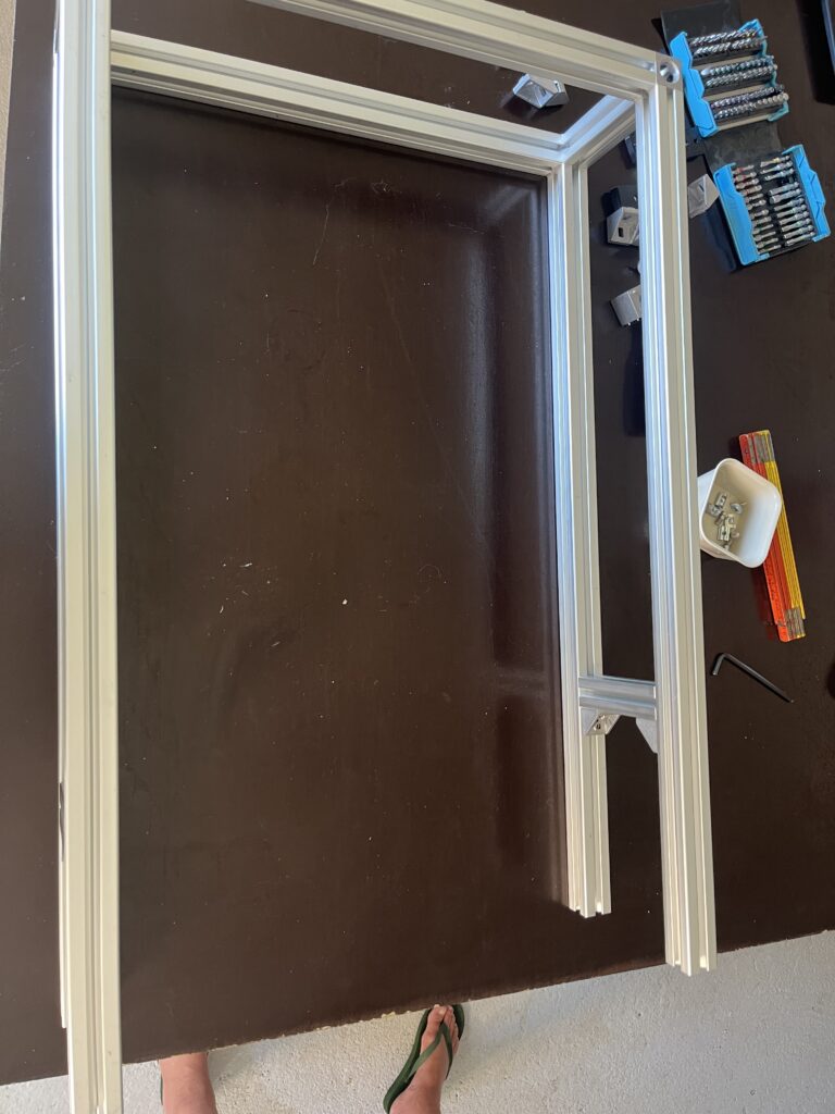 Aluminum frame sections laid out on a dark worktable with a blue tool case and small hardware nearby; a person’s feet in green flip-flops are visible at the bottom.