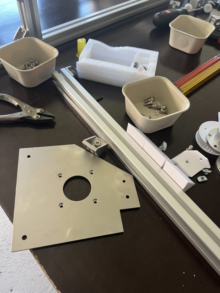 Workbench with metal mounting plate, aluminum extrusion rail, bins of screws and bolts, and a foam packing block on the table.
