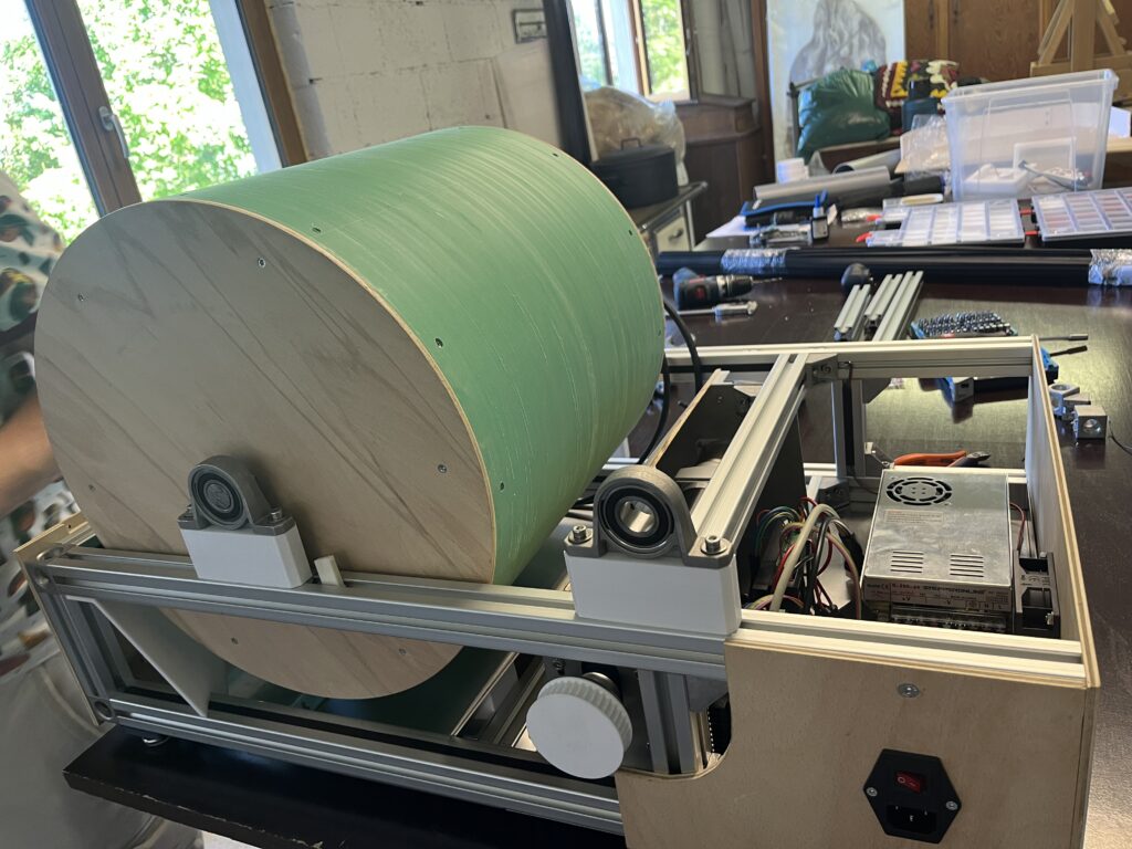 Large green cylindrical drum mounted on a metal machine in a workshop, with tools and wiring visible nearby as background.