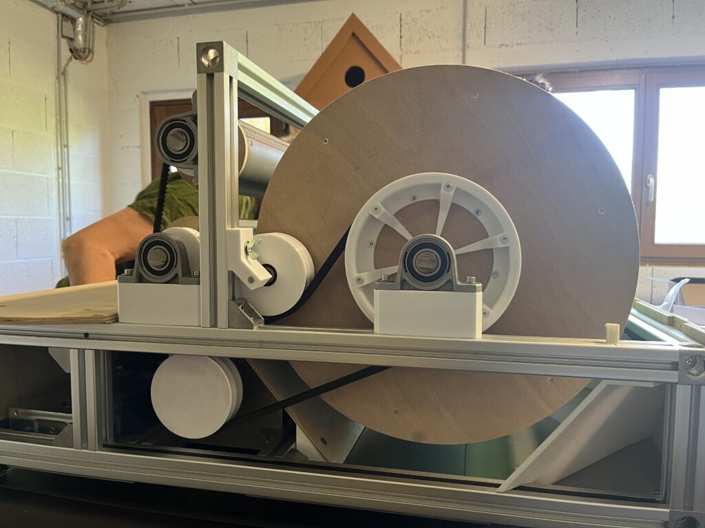 Industrial packaging/printing machine with a large brown spool, white rollers, and aluminum frame in a workshop setting