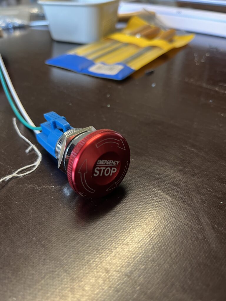 Red emergency stop button mounted on blue hardware, wires nearby on a dark work surface with blurred packaging in the background.