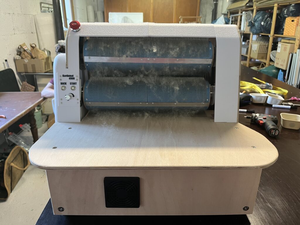 Front view of a Kardomat 4900 industrial roller machine in a workshop, three blue cylindrical rollers with dust and fibers on them.